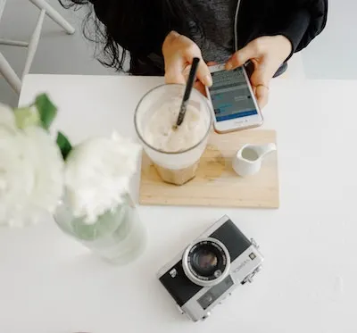 a white table with someone uses a phone, coffee, flowers and a camera