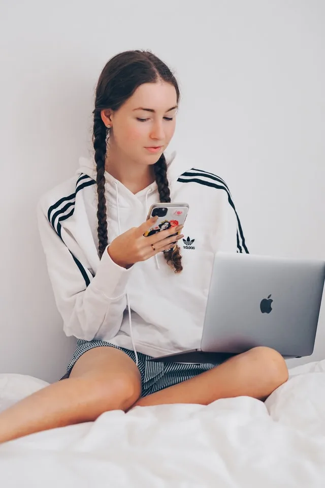 girl sitting on a bed with laptop on the lap and phone in the hand. Blog graphic on How to make money as a teenager without a job