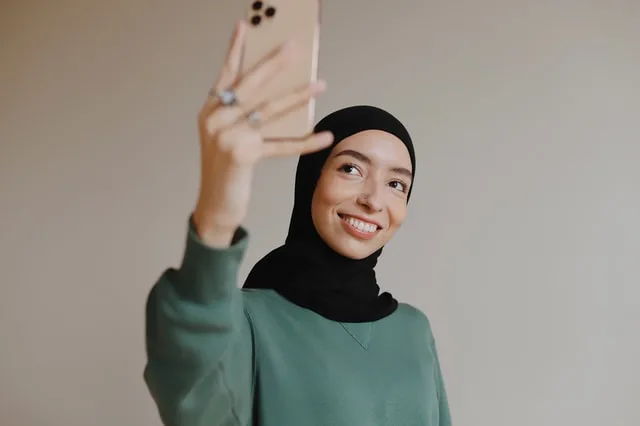 a girl with a hijab, holding an iphone and taking a selfie. Graphic for a post on how much do tiktokers make money