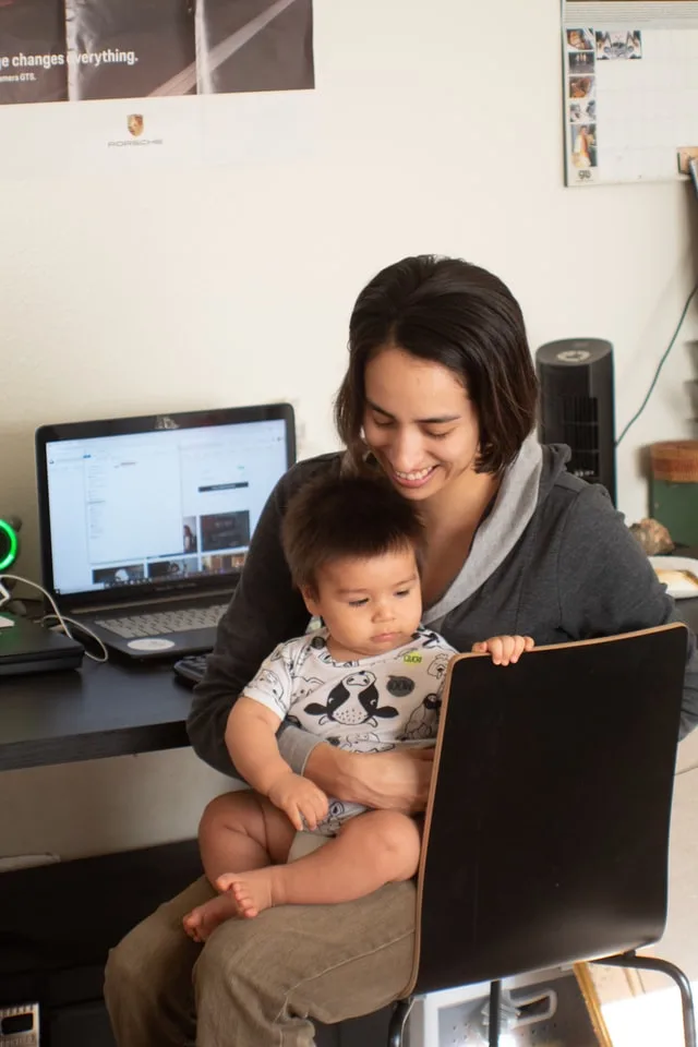 In this image a mother is sitting on a chair near her workstation with along with her small kid sitting on her lap. Graphic is used for a blog post on jobs for homemaker