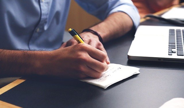 A table with laptop, hands writing on a notepad with a pen.