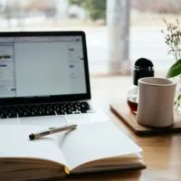 This image includes a laptop, notebook, pen, coffe mug, flowers in a vase
