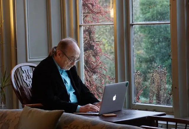 In this image an old man is working on a laptop from the comfort of his home