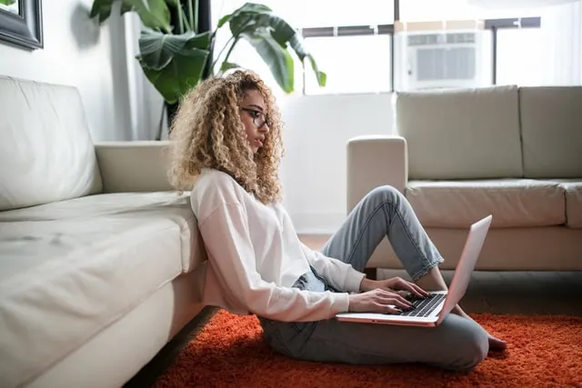 In  this image a women is sitting on the floor leaning backwards towards her sofa while working on her laptop, Image for a post on how to make money fast as a woman