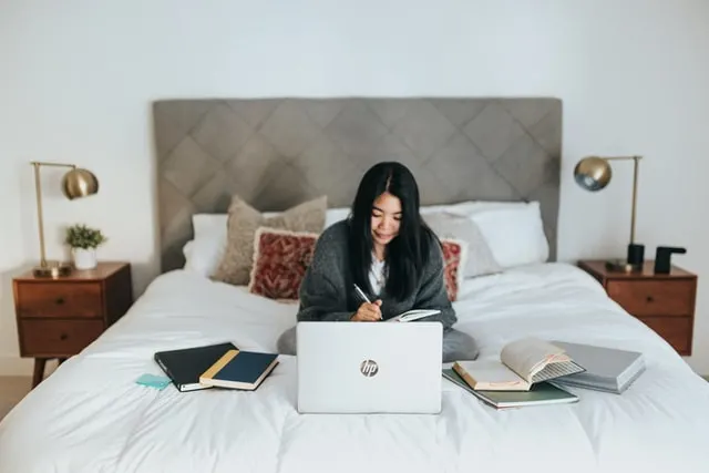 In this image a women is sitting on her bed. She is making notes while watching something on her laptop. There are few books kept on her both sides on the bed.