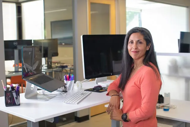 In this image a women confidently showcasing her work table which consists of a desktop, a laptop, and some stationary