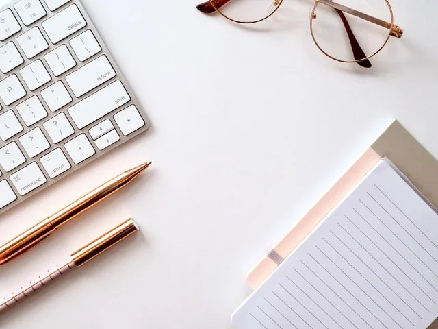 In this image a keyboard, few pens, one pair of spectacles, notepads are kept on a workstation