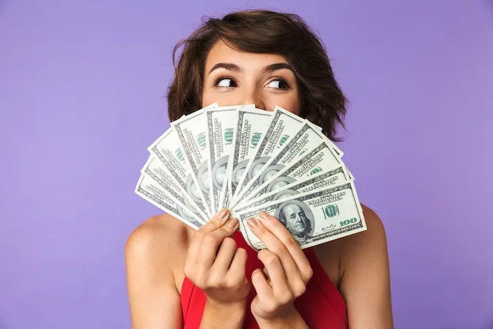 Happy Pretty brunette woman covering her face with money and looking away over purple background