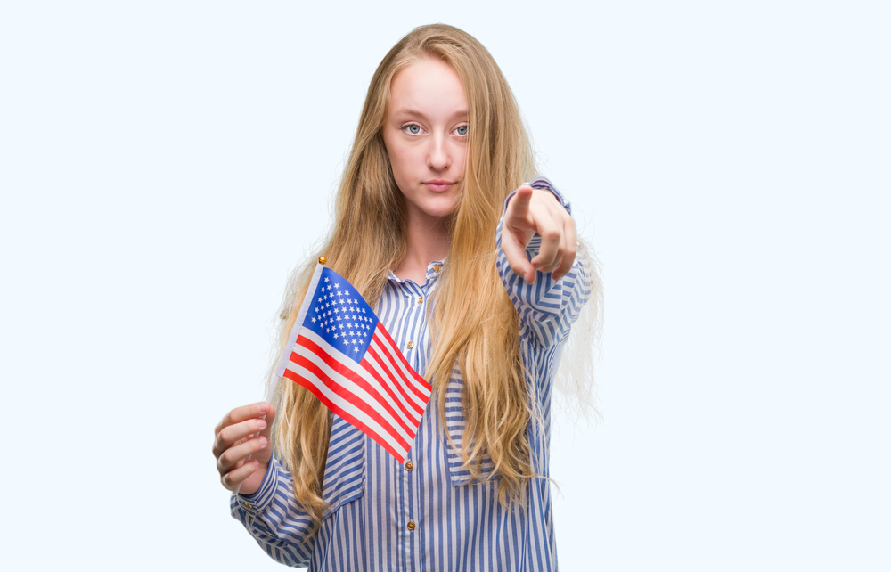 Blonde teenager woman holding flag of United States of America pointing with finger to the camera and to you, hand sign, positive and confident gesture from the front