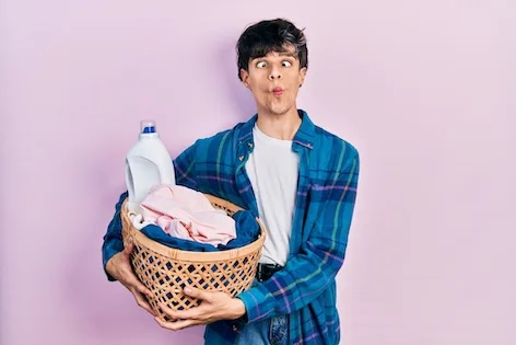 Handsome hipster young man holding laundry basket and detergent bottle making fish face with mouth and squinting eyes, crazy and comical.