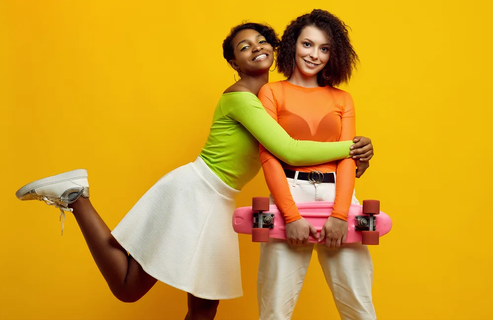 Portrait of two young beautiful hipster girls in trendy colorful summer clothes on yellow background. Crazy positive bright teens