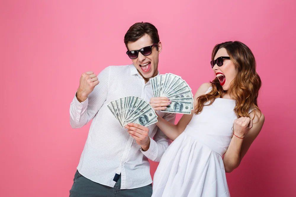 Portrait of a satisfied smartly dressed couple holding bunch of money banknotes and celebrating isolated over pink background
