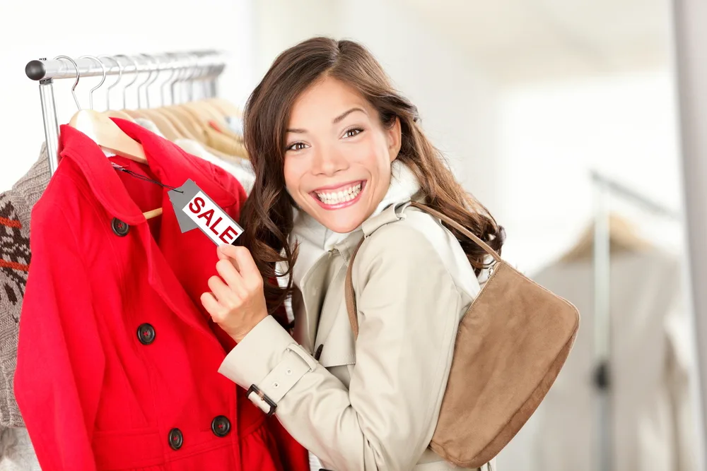 Shopping woman excited showing price tag at clothes sale in clothing store. Smiling cheerful woman. Price label reads sale.