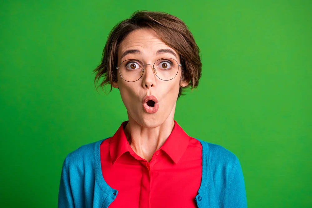 Portrait of young beautiful shocked amazed impressed speechless girl wear glasses isolated on green color background.