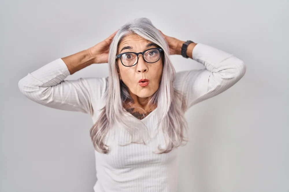 Middle age woman with grey hair standing over white background crazy and scared with hands on head, afraid and surprised of shock with open mouth