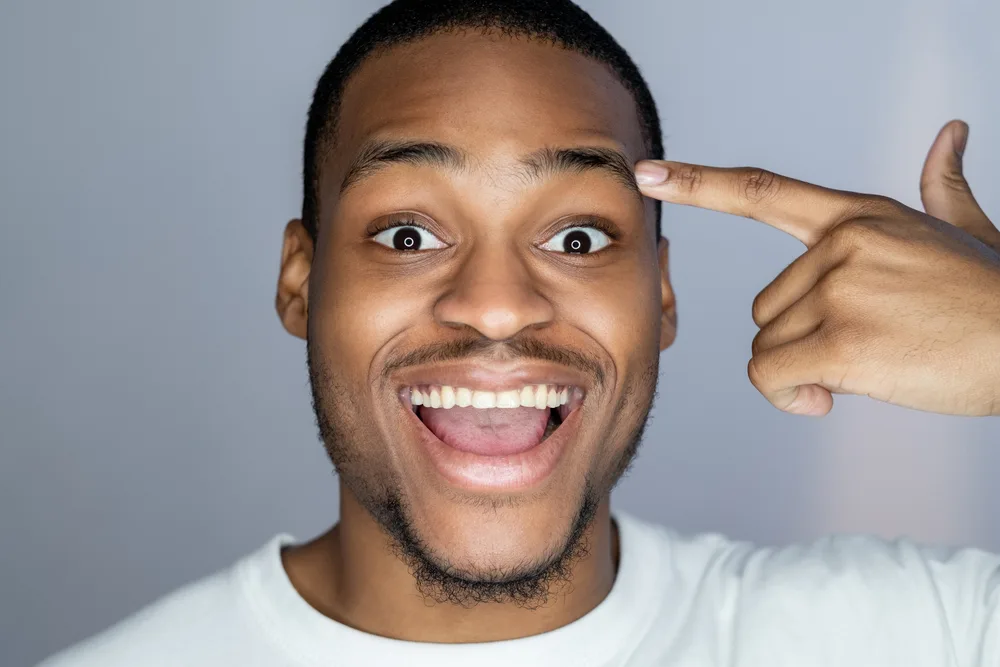 Enthusiastic African man. Aha moment. Self motivation. Portrait of happy astonished handsome dark skin guy in white pointing at face temple smiling isolated on gray background.