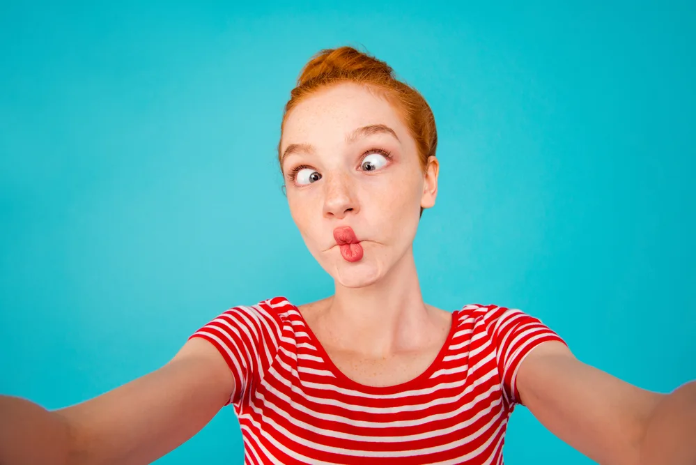 Self-portrait of crazy foolish playful pretty ecstatic cheerful red-haired girl with bun, grimacing, isolated on bright vivid blue background