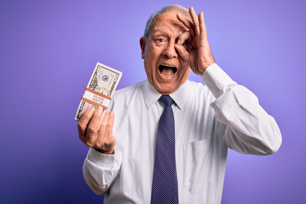 Senior grey haired man holding bunch of fifty dollars banknotes over purple background with happy face smiling doing ok sign with hand on eye looking through fingers