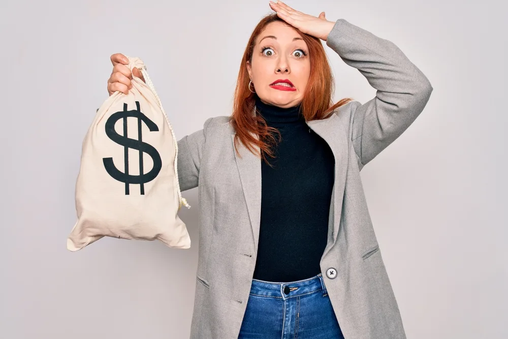 Young beautiful redhead woman holding bag with money and dollar sign over white background stressed with hand on head, shocked with shame and surprise face, angry and frustrated. Fear and upset for mistake.