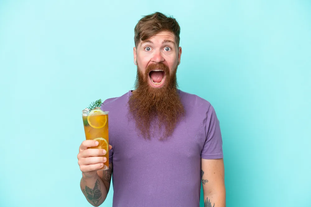 Redhead man with long beard holding a cocktail isolated on blue background with surprise and shocked facial expression