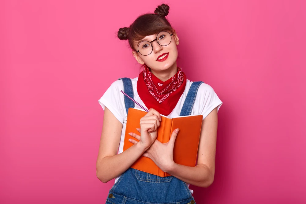 Studio image of cute female teenager with two funny bunches, holding diary and pensil in hands, looking directly at camera, writes secrets in her orange notebook. People, youth, lifestyle concept.