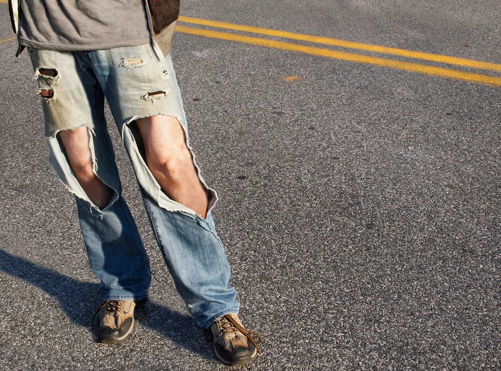 A young man, from the waist down, in a causal t-shirt and ripped jeans with hair legs exposed stands in the road.