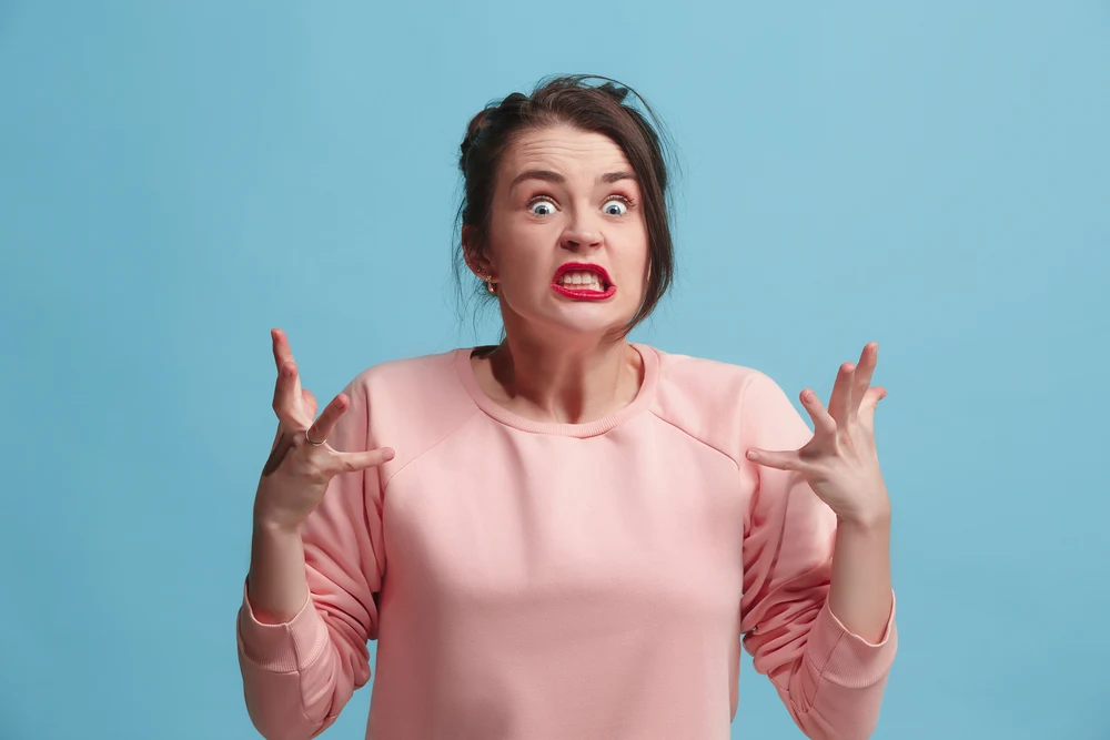 Angry woman looking at camera. Aggressive business woman standing isolated on trendy blue studio background. Female half-length portrait. Human emotions, facial expression concept. Front view.