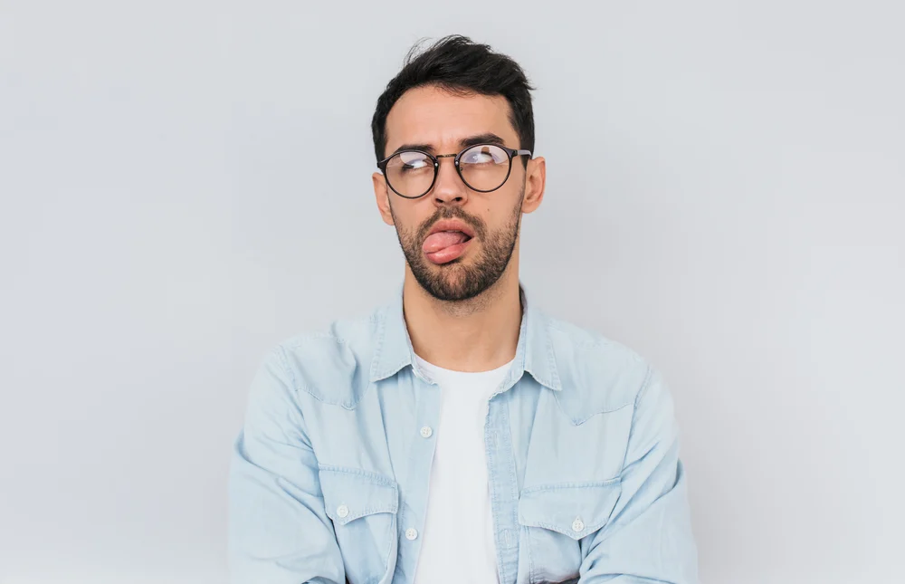 Portrait of stylish bearded male wears round trendy glasses and blue shirt, has disgusted expression with tongue. Human facial expressions, emotions, people and feelings concept