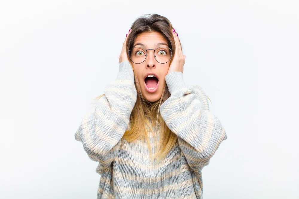 young pretty woman looking unpleasantly shocked, scared or worried, mouth wide open and covering both ears with hands against white background