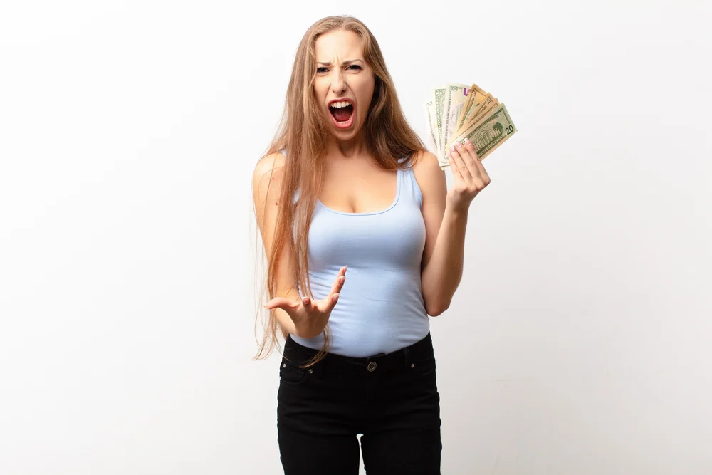 yound blonde woman looking angry, annoyed and frustrated screaming wtf or what’s wrong with you holding dollar banknotes