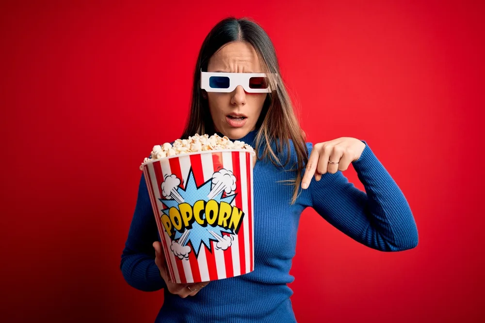 Young blonde woman wearing 3d glasses and eating pack of popcorn watching a movie on cinema Pointing down with fingers showing advertisement, surprised face and open mouth