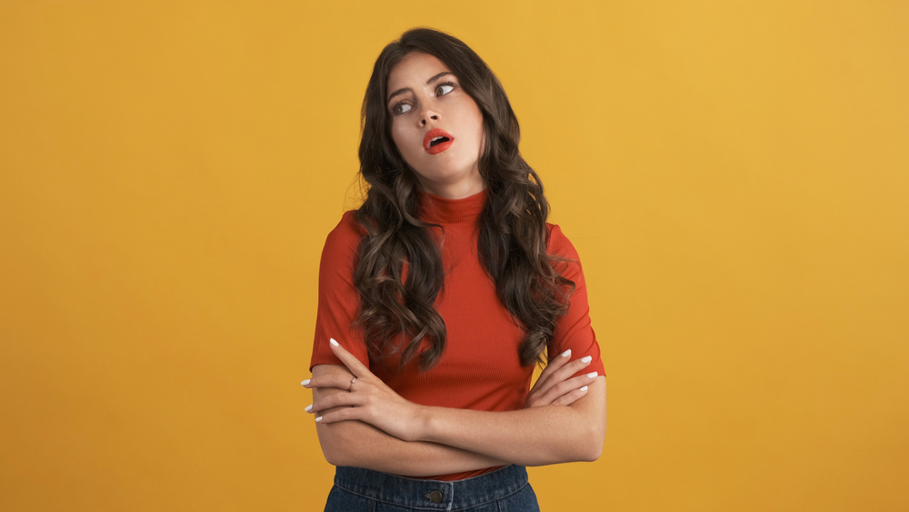 Casual bored brunette girl in red top with hands crossed tiredly posing on camera over yellow background. Not interested expression