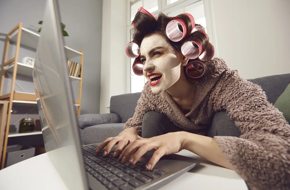 Mad woman in face mask and hair rollers sitting looking at laptop computer screen, writing insulting hater comment, spreading fake rumour. Jealous wife reading husband's direct message on social media