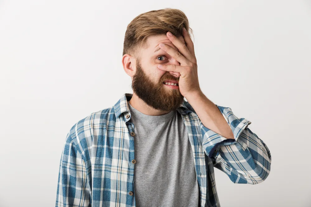 Ashamed bearded man dressed in plaid shirt standing isolated over white background, cover face