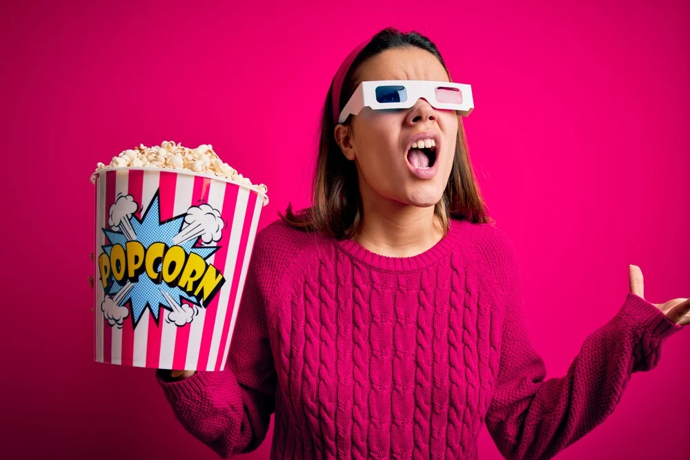 Young beautiful girl watching movie using 3d glasses eating box with popcorns crazy and mad shouting and yelling with aggressive expression and arms raised. Frustration concept.