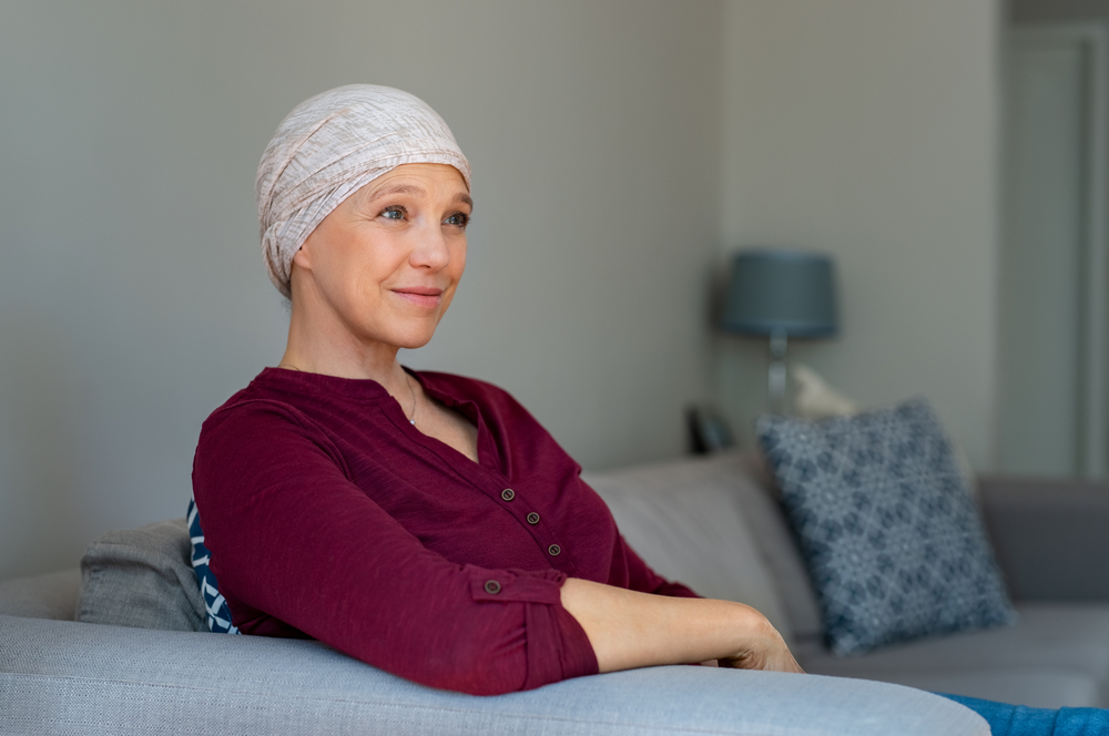 Portrait of mature woman recovering after chemotherapy. Senior woman fighting breast cancer and wearing a headscarf. Hopeful ill lady at home covering head with scarf looking away while sitting on sofa.