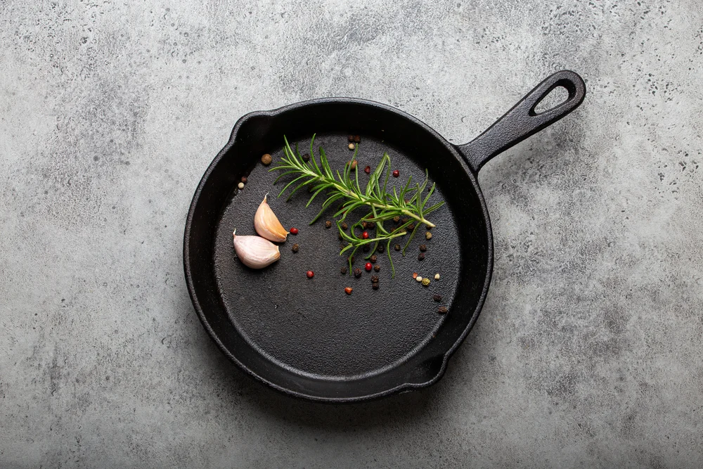 Black cast iron frying pan with ingredients for cooking: fresh green rosemary, raw garlic cloves and colorful peppercorns on gray stone background flat lay top view, cooking food concept