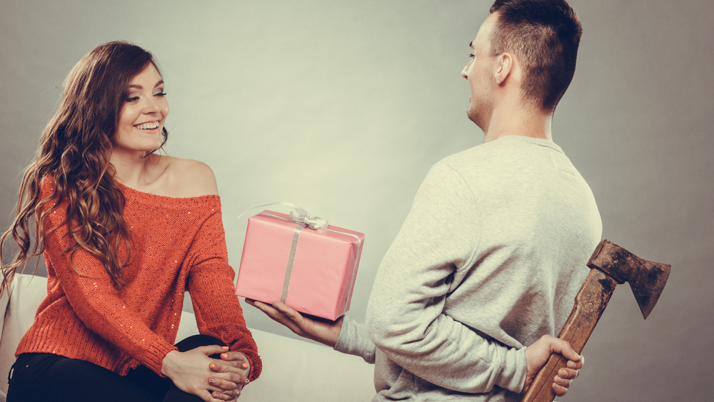 Sneaky insincere man holding axe giving gift present box to woman. Husband concealing hiding his true feelings from happy trusting wife. Untrue false intention. Relationship problems. Instagram.