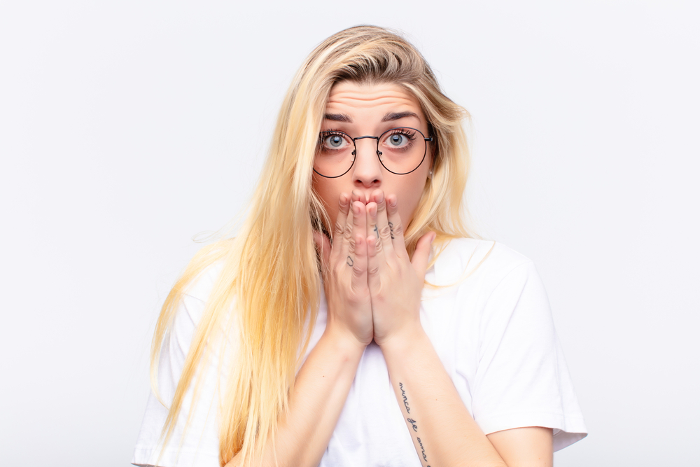 young pretty blonde woman feeling worried, upset and scared, covering mouth with hands, looking anxious and having messed up against white wall