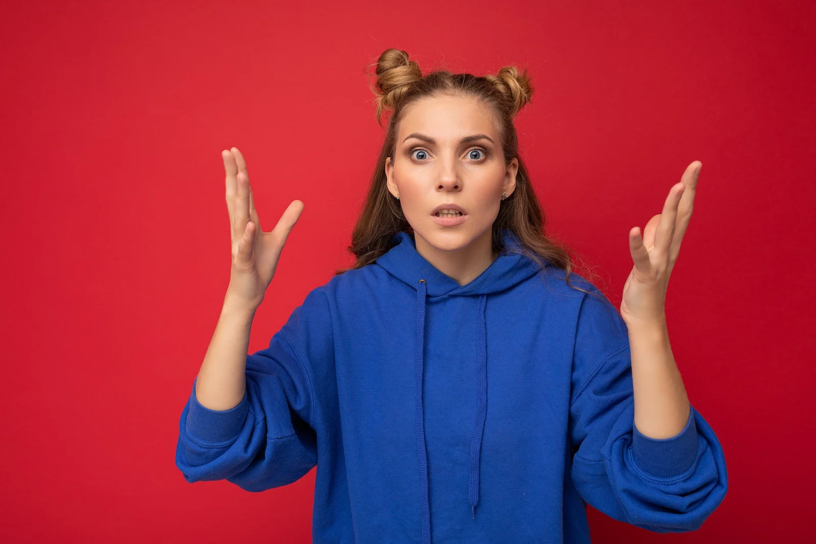 Photo of shocked emotional young beautiful blonde woman with two horns with sincere emotions wearing hipster bright blue hoodie isolated over red background with free space. Wow concept