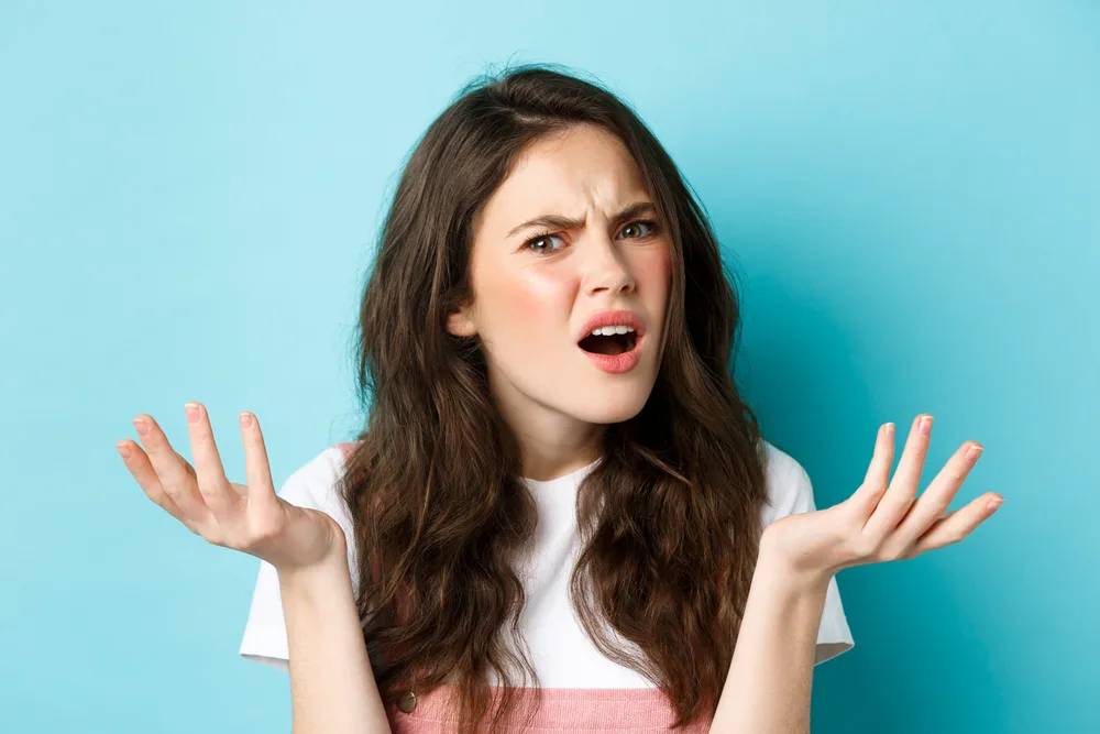 Image of confused and annoyed young woman shrugging, asking what is that, staring questioned and displeased at camera, standing against blue background.