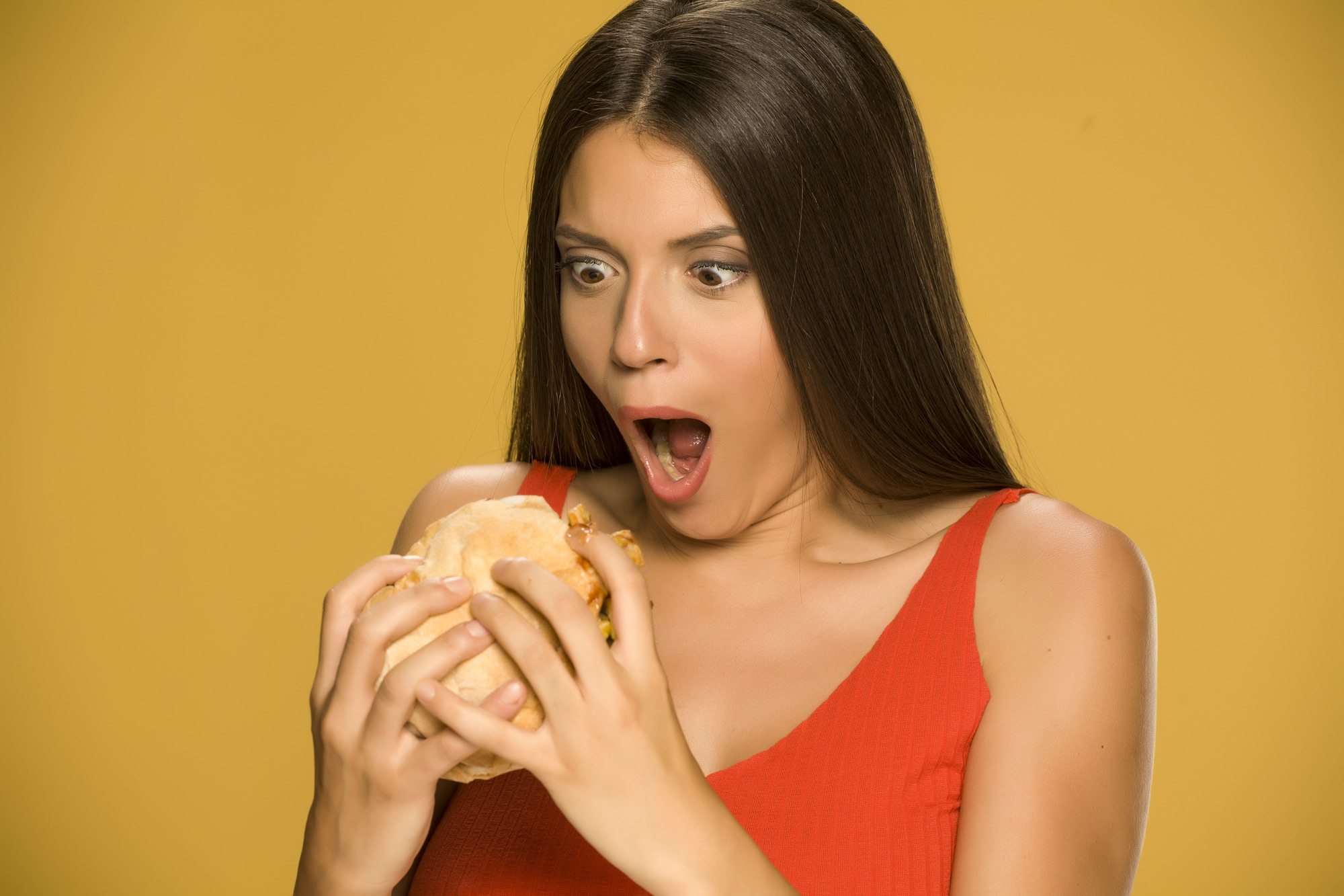 Young greedy woman eating a burger on yellow background