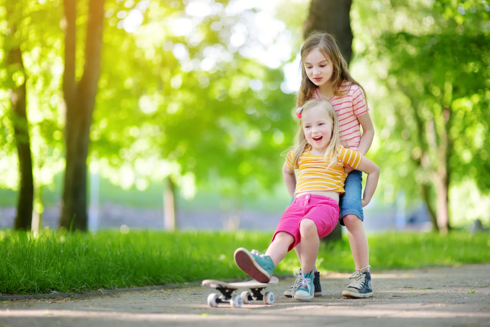 Two pretty little girls learning to skateboard on beautiful summer day in a park. Children enjoying skateboarding ride outdoors.