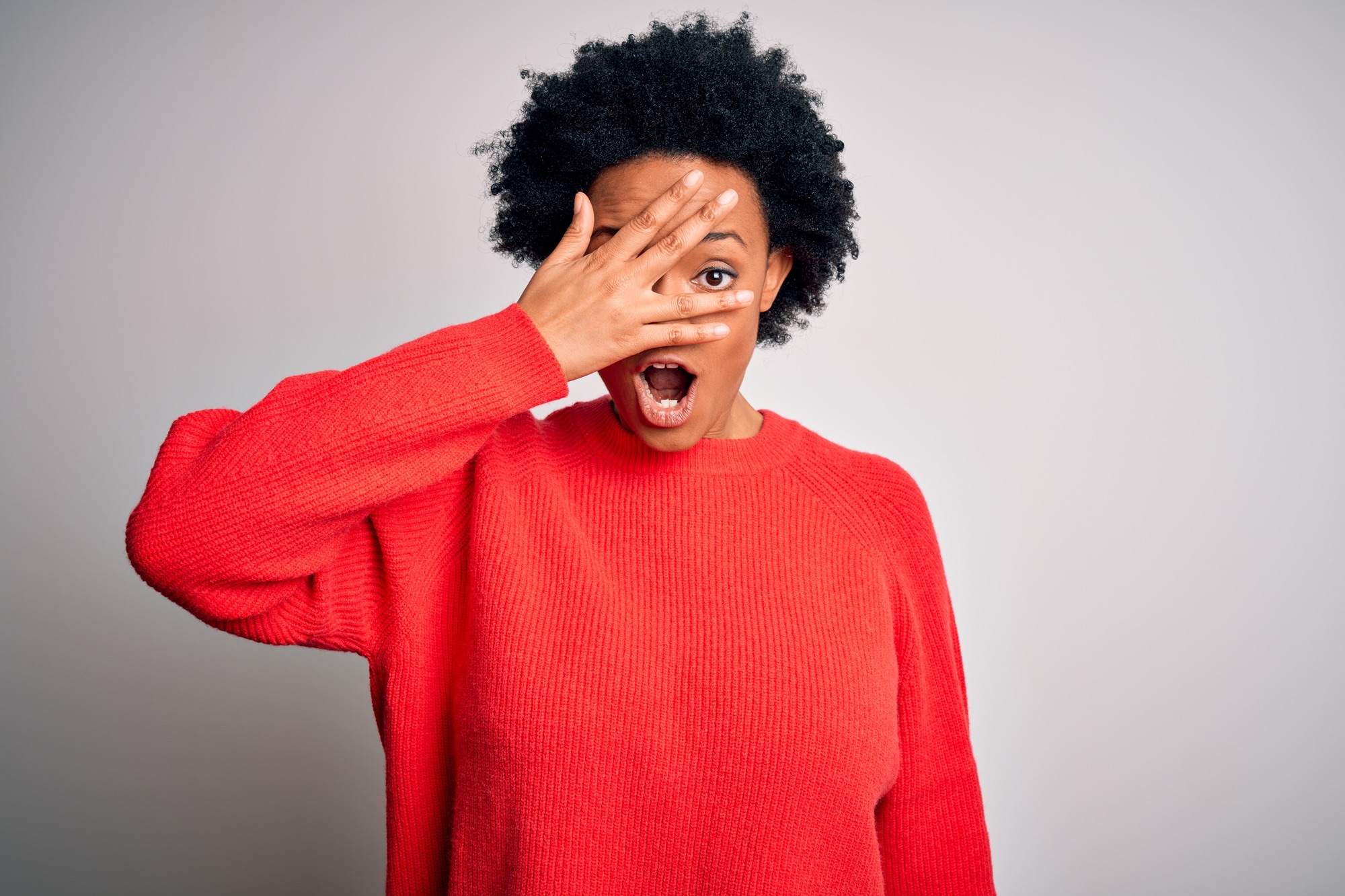 Young beautiful African American afro woman with curly hair wearing red casual sweater peeking in shock covering face and eyes with hand, looking through fingers with embarrassed expression.