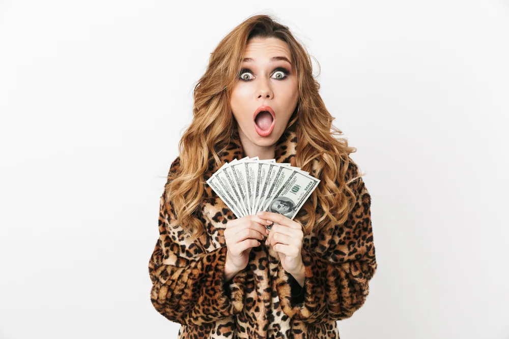 Beautiful young woman wearing leopard coat standing isolated over white background, showing money banknotes