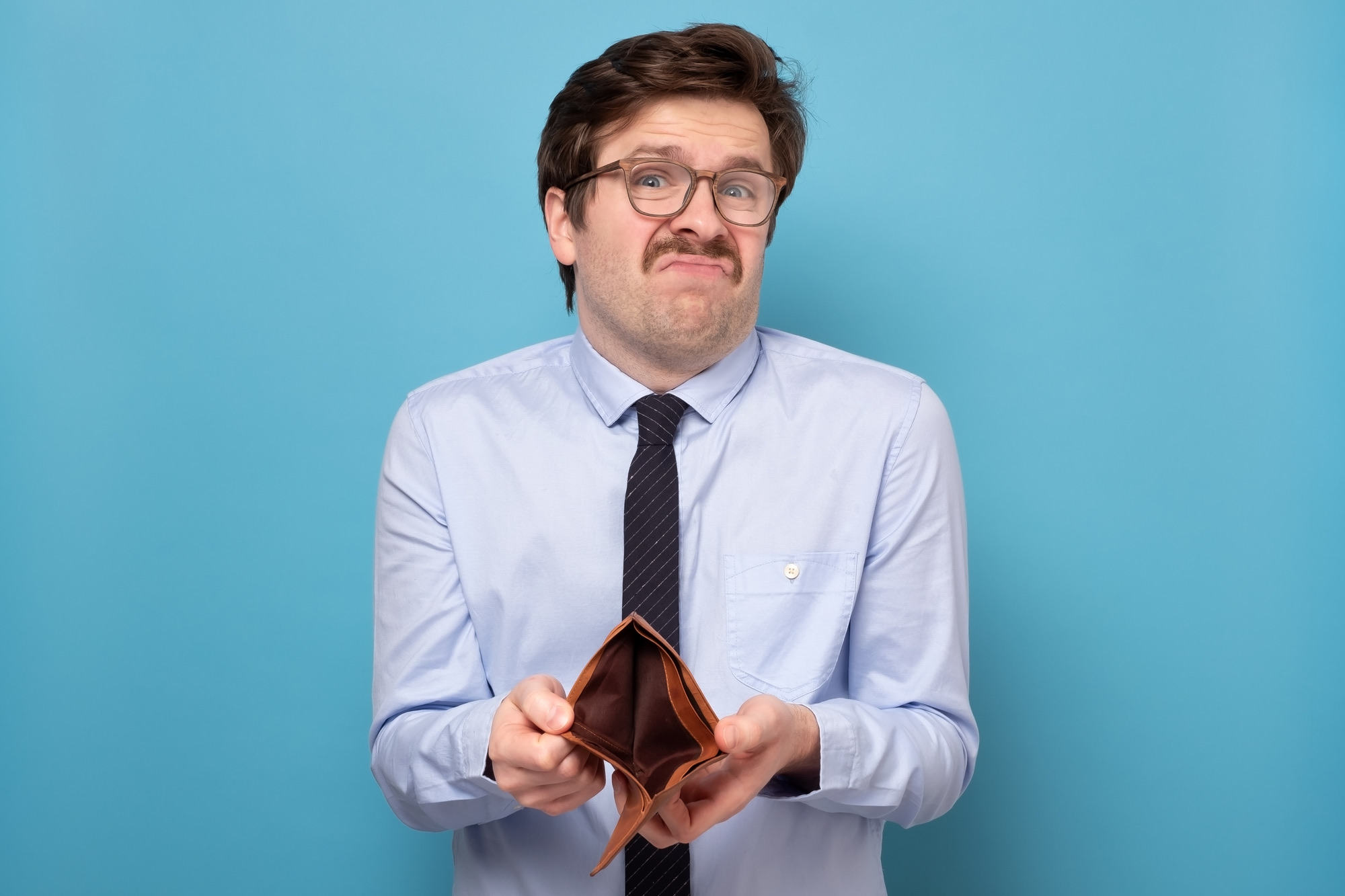 Sad businessman holding an empty wallet isolated on a blue background. Bankruptcy financial difficulties concept. Job loss.