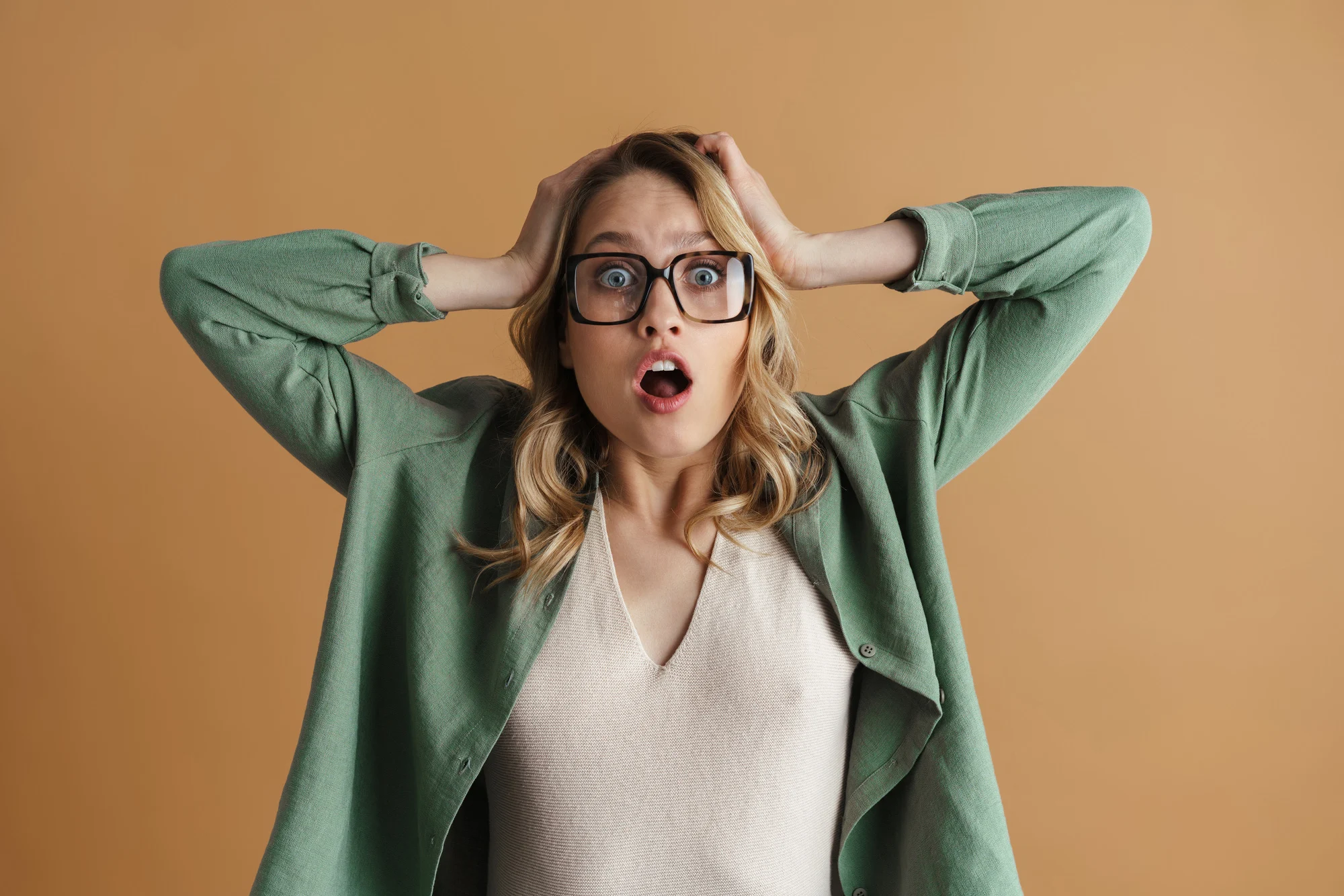 Shocked beautiful woman in eyeglasses grabbing her head isolated over beige wall