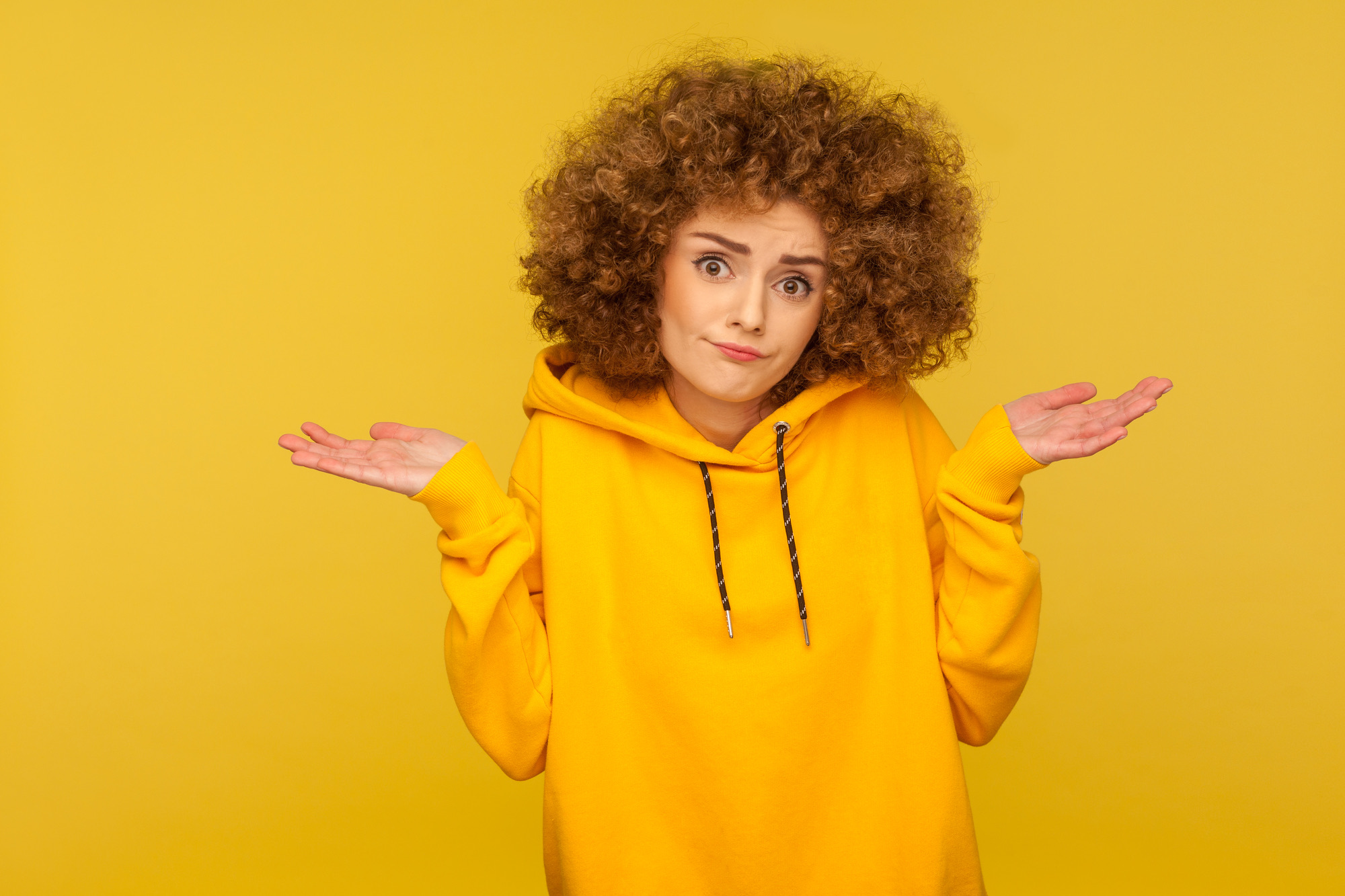 Don't know, sorry. Portrait of clueless uncertain confused curly-haired woman in urban style hoodie shrugging shoulders in questioning gesture, looking with indifference. indoor studio shot isolated