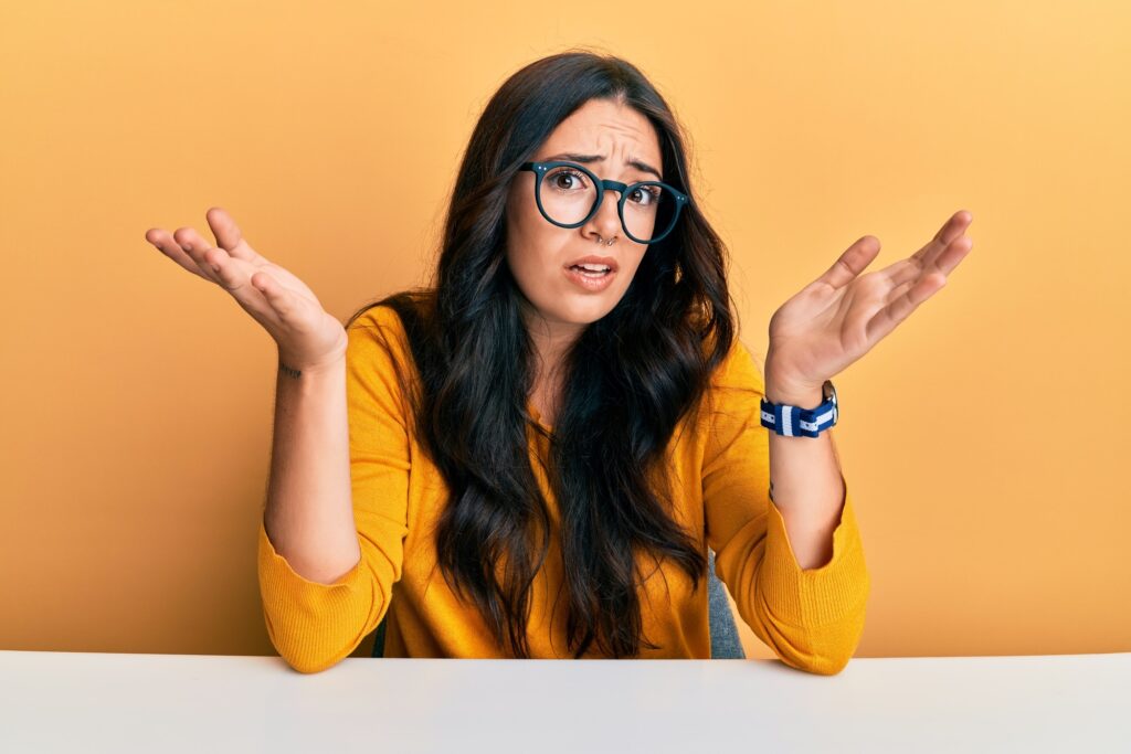 Beautiful brunette young woman wearing glasses and casual clothes sitting on the table clueless and confused expression with arms and hands raised. doubt concept.