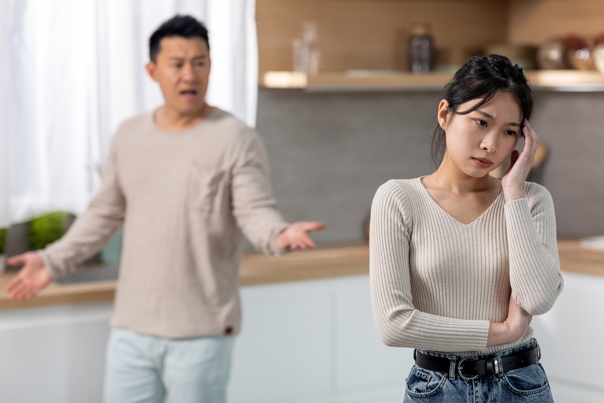 Upset young asian woman touching her head, thinking about divorce, angry middle aged man husband shouting at his wife, kitchen interior. Domestic abuse, violence in marriage concept